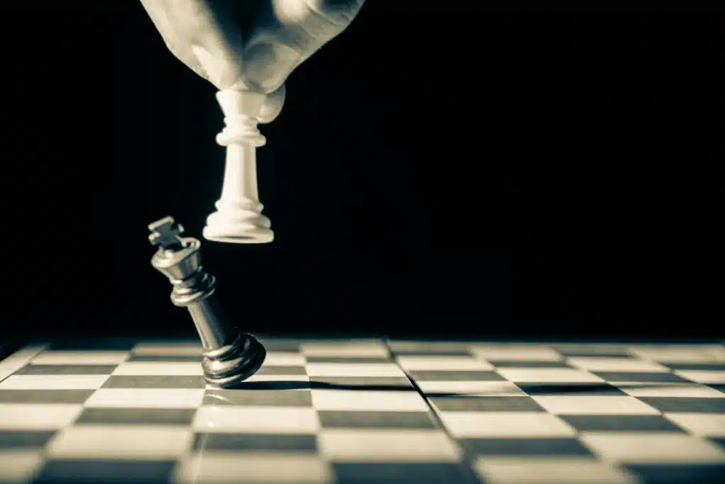 Das Bild zeigt eine Schachszene mit einer Hand, die eine weiße Schachfigur bewegt, während eine schwarze Figur kippt. Die Darstellung symbolisiert Strategie, Wettbewerb, taktische Entscheidungen und den intelligenten Umgang mit Herausforderungen im Zusammenhang mit EWM AG.